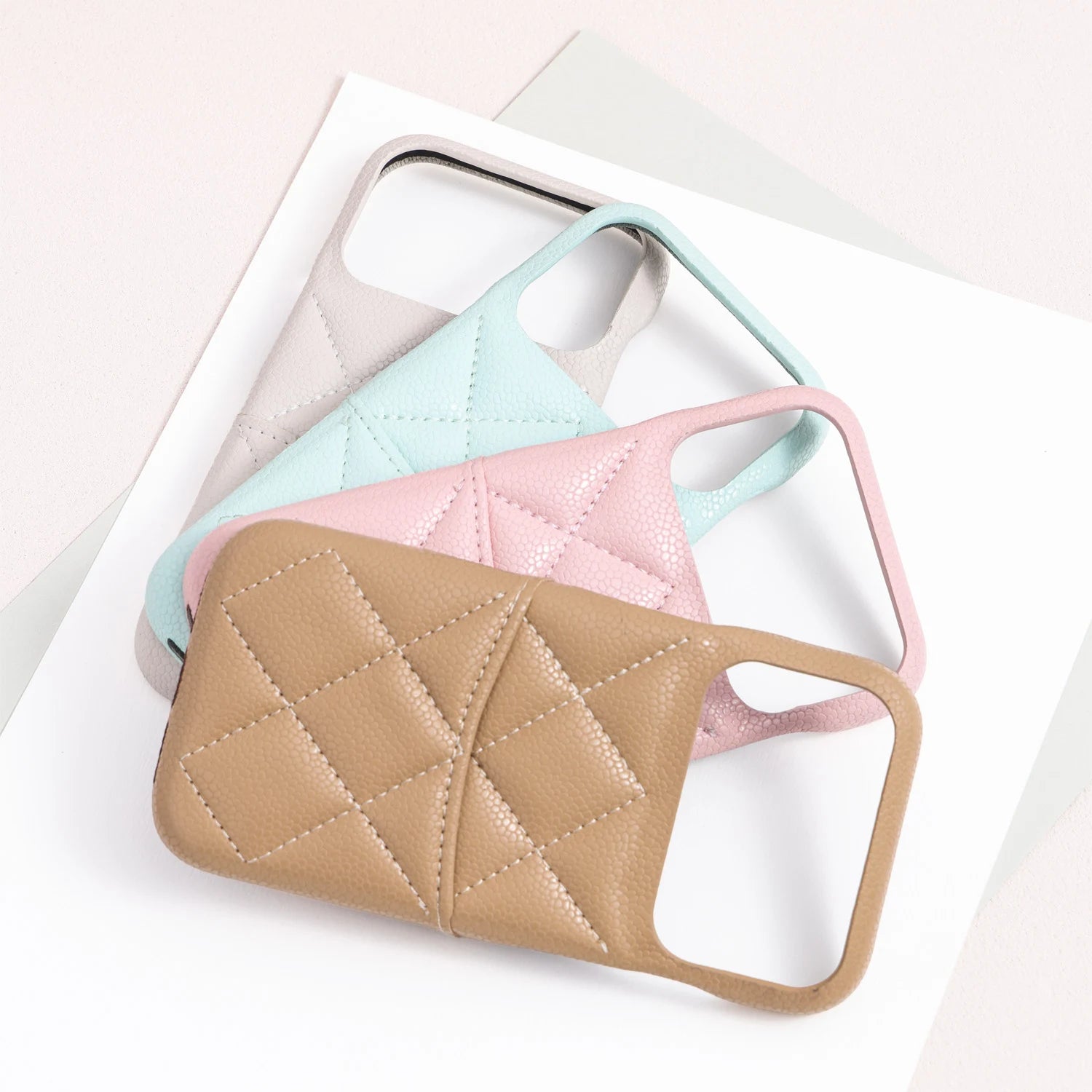 Overlapping textured leather phone cases in ivory, sky blue, powder pink, and beige showing the geometric quilted stitch pattern.