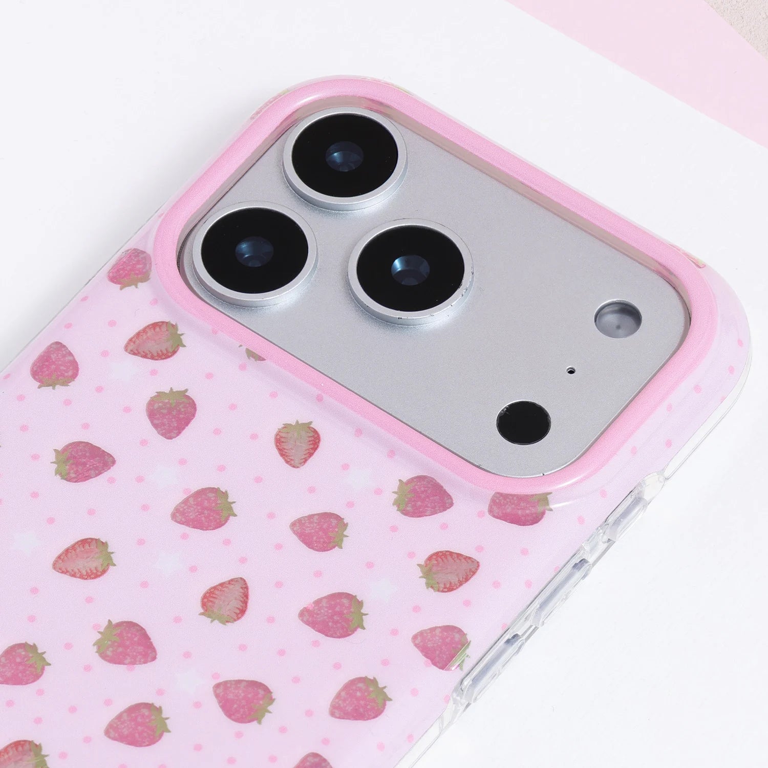 Close-up of a soft pink strawberry phone case with miniature watercolor-style strawberries, tiny tonal dots, and a pink camera surround.