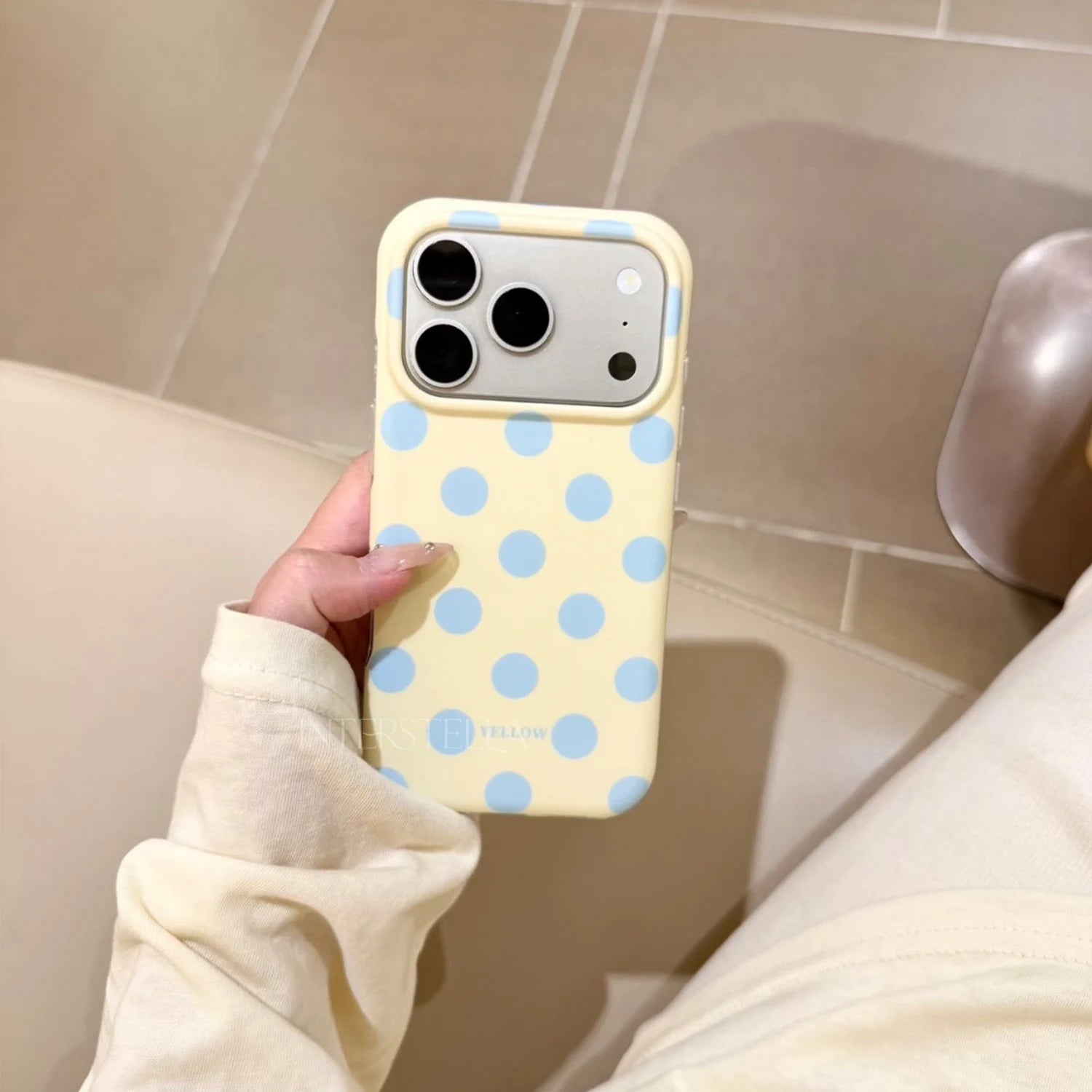 Hand holding a butter yellow phone case with powder blue polka dots and raised camera surround in a soft indoor setting.