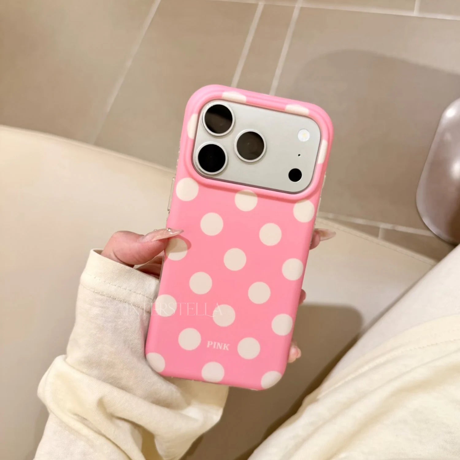 Hand holding a candy pink phone case with ivory polka dots and raised camera surround in a warm indoor setting.