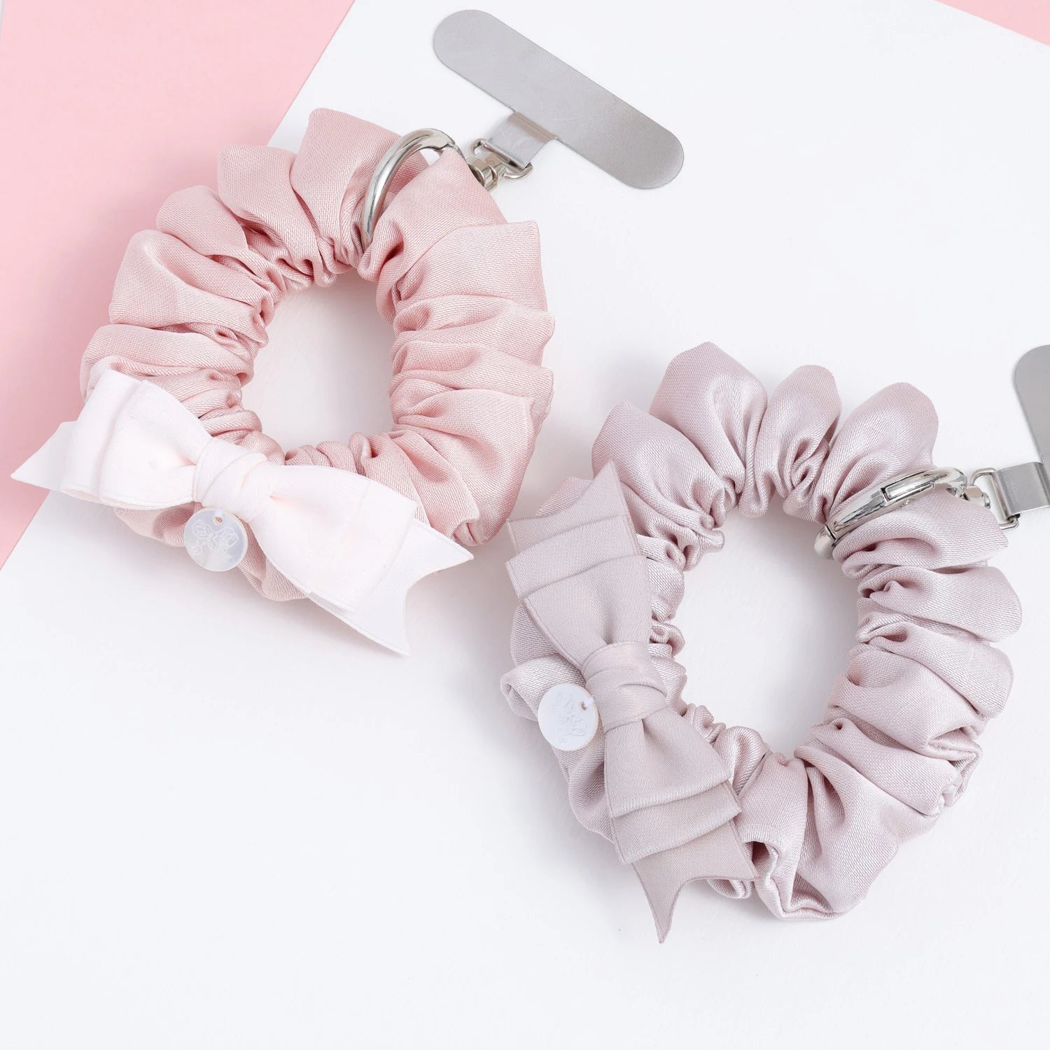 Two fabric phone hanging rings shown together, each with a gathered scrunchie silhouette, decorative bow, and polished metal attachment hardware in soft pink colorways.
