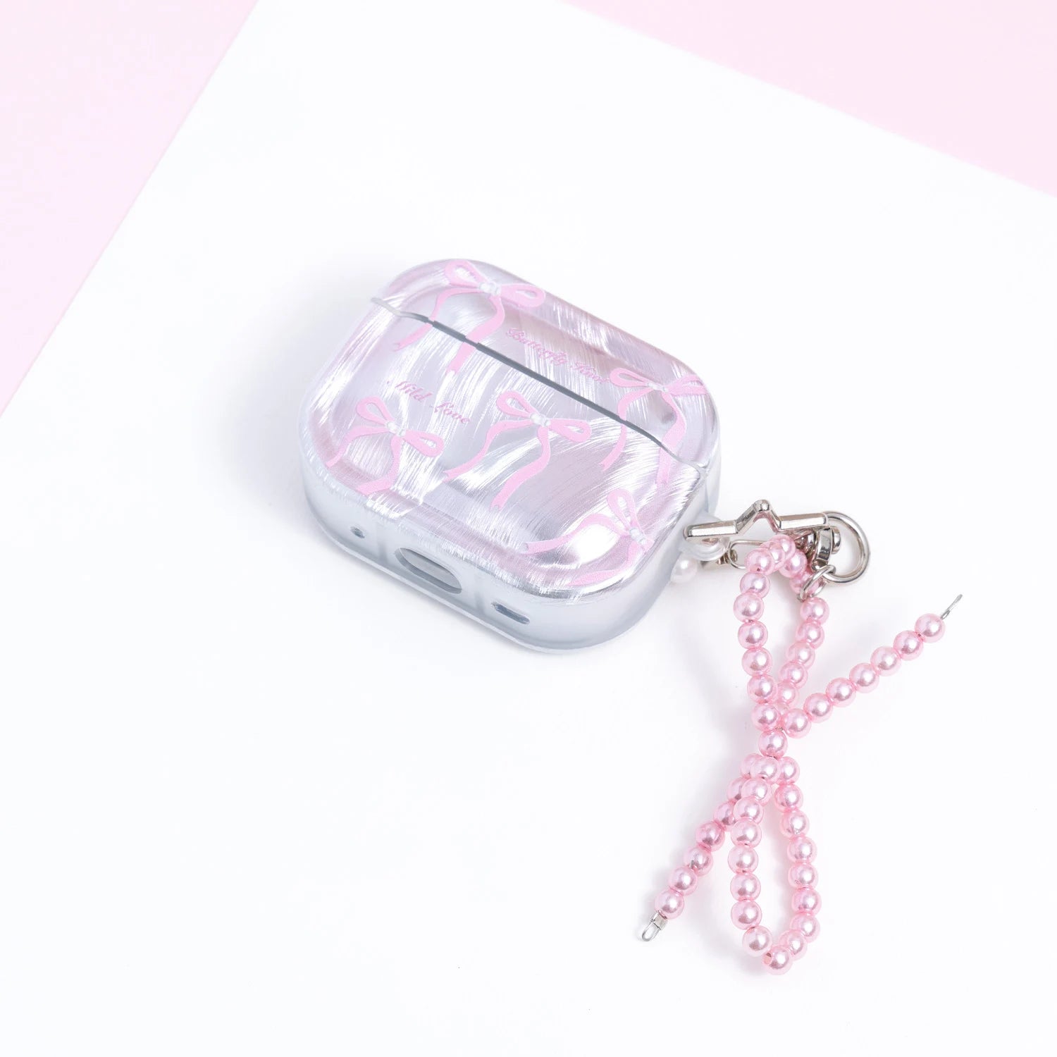 Closed pearly silver AirPods case with pink ribbon print and a dangling pink pearl-bead bow charm displayed on a soft pink-and-white background.