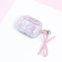 Closed pearly silver AirPods case with pink ribbon print and a dangling pink pearl-bead bow charm displayed on a soft pink-and-white background.
