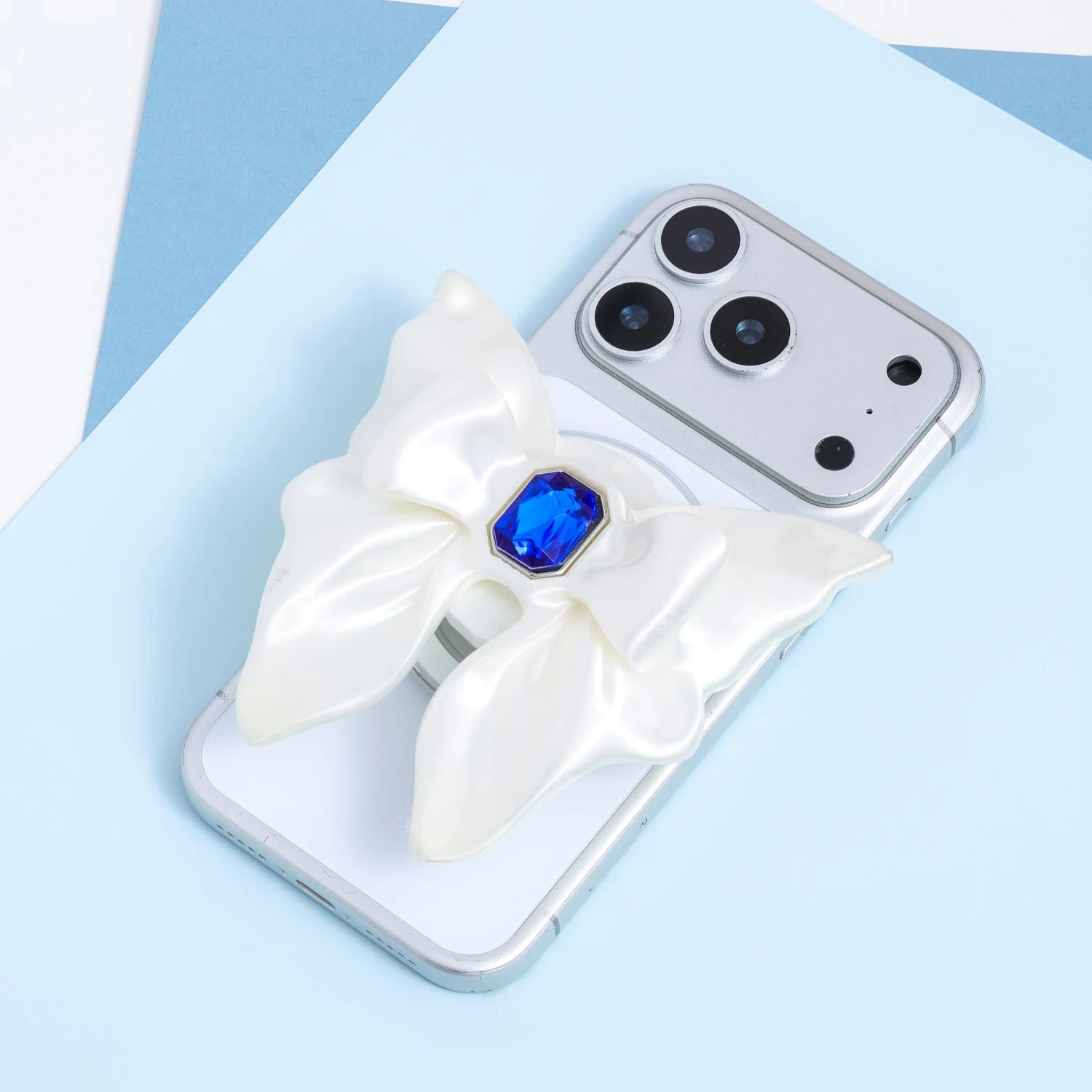 A silver phone fitted with a self-designed 3D-printed ivory bow phone stand, featuring a sculptural ribbon shape and an embedded sapphire-blue jewel at the center.