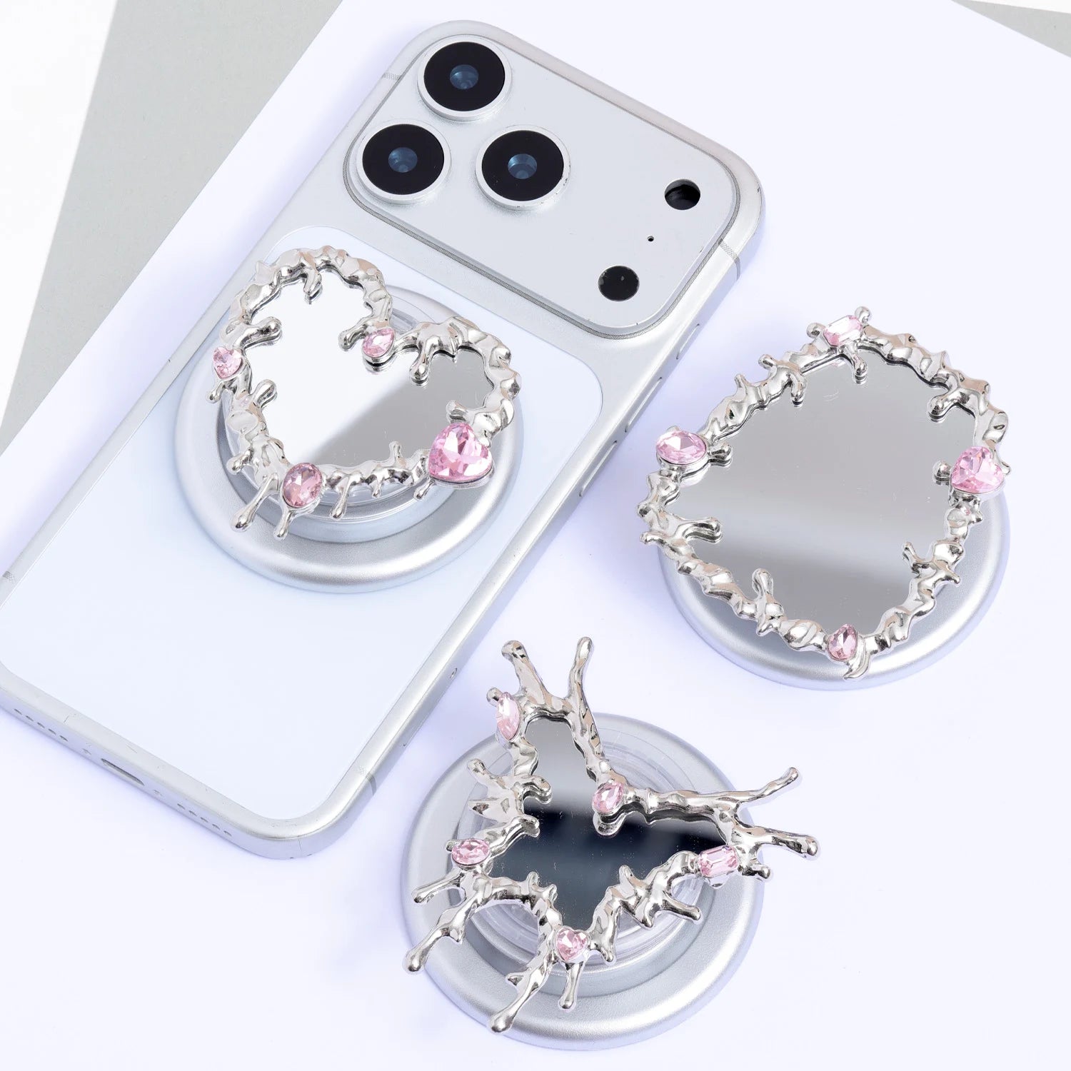 Three silver-tone mirror phone stands with pink crystal accents displayed together, featuring heart, cross-like, and ornamental frame silhouettes on circular bases.