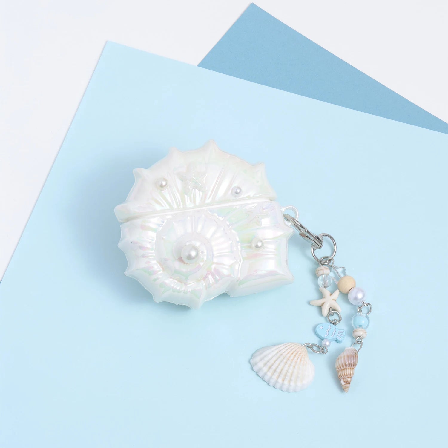 Closed iridescent conch-shaped AirPods case in opaline pearl white with embossed shell detailing and a dangling ocean charm chain with shells, beads, and starfish accents.