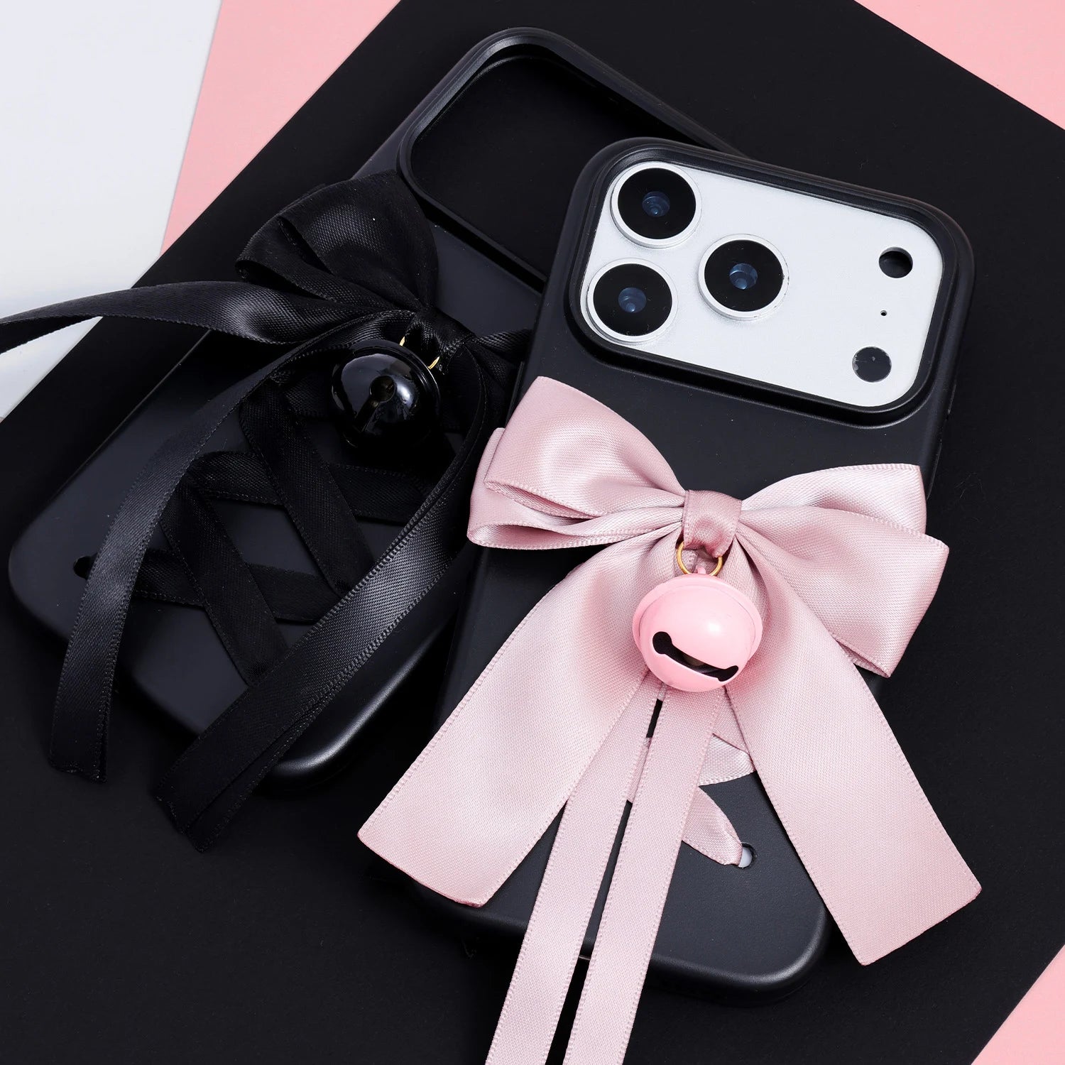 Two matte black phone cases displayed together, one with a blush pink satin bow and matching bell charm, and one with a black satin bow and black bell charm.
