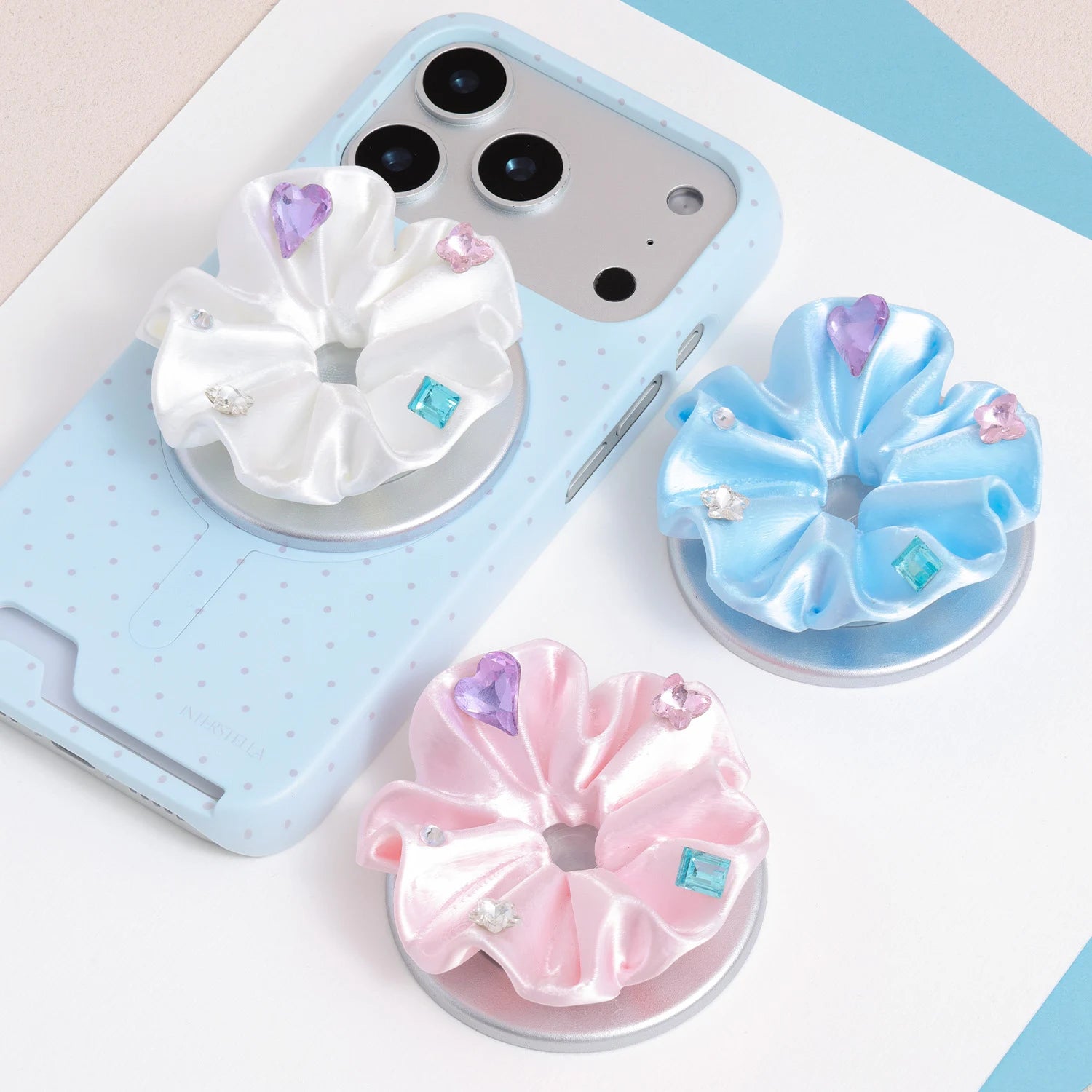 Three pastel jewel rosette phone grip stands in white, pink, and blue displayed beside a light blue polka dot card-slot phone case.