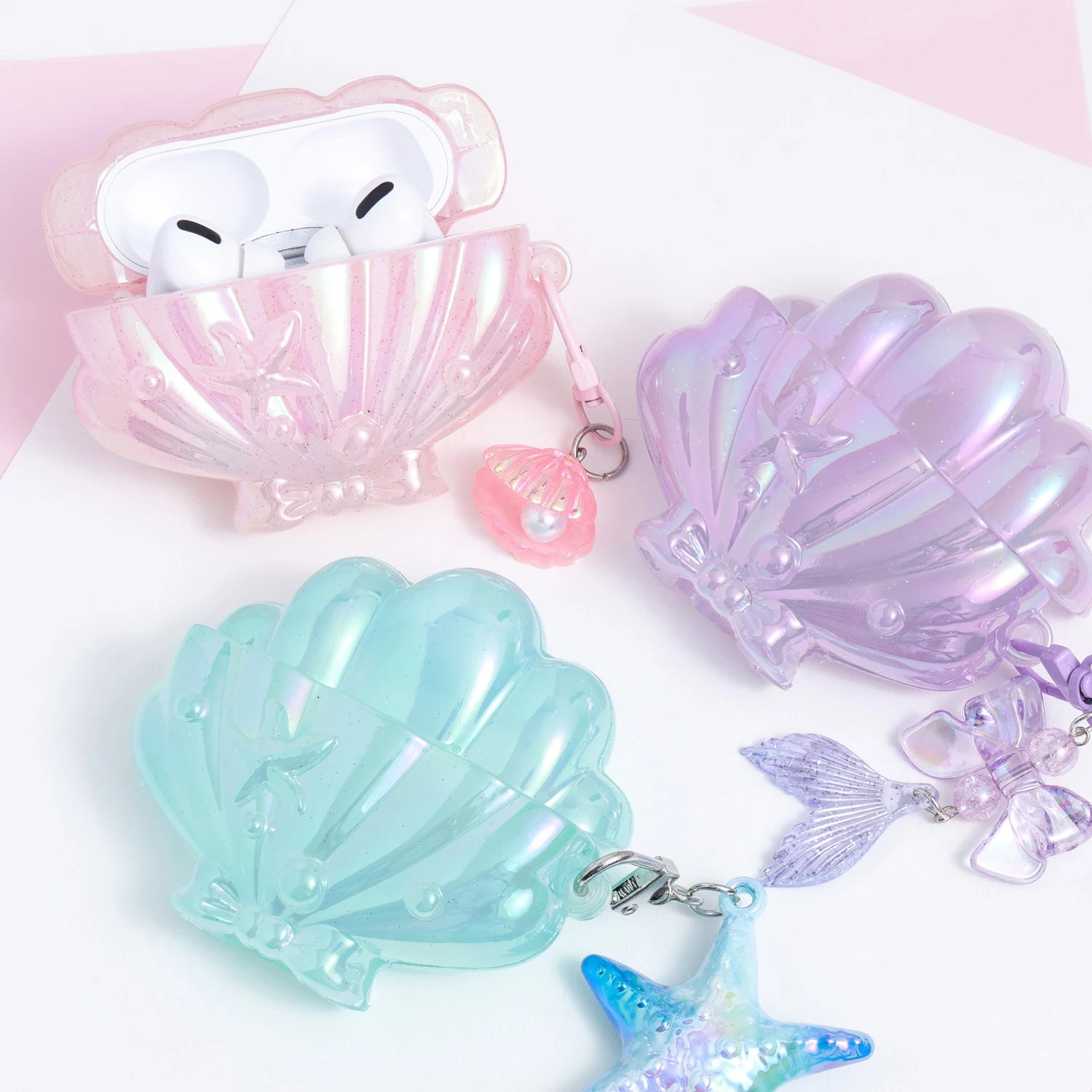 Three iridescent shell-shaped AirPods cases in pink, lilac, and aqua pastel tones, each finished with an ocean-inspired dangling charm including a shell pearl, mermaid tail, and starfish.