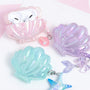 Three iridescent shell-shaped AirPods cases in pink, lilac, and aqua pastel tones, each finished with an ocean-inspired dangling charm including a shell pearl, mermaid tail, and starfish.