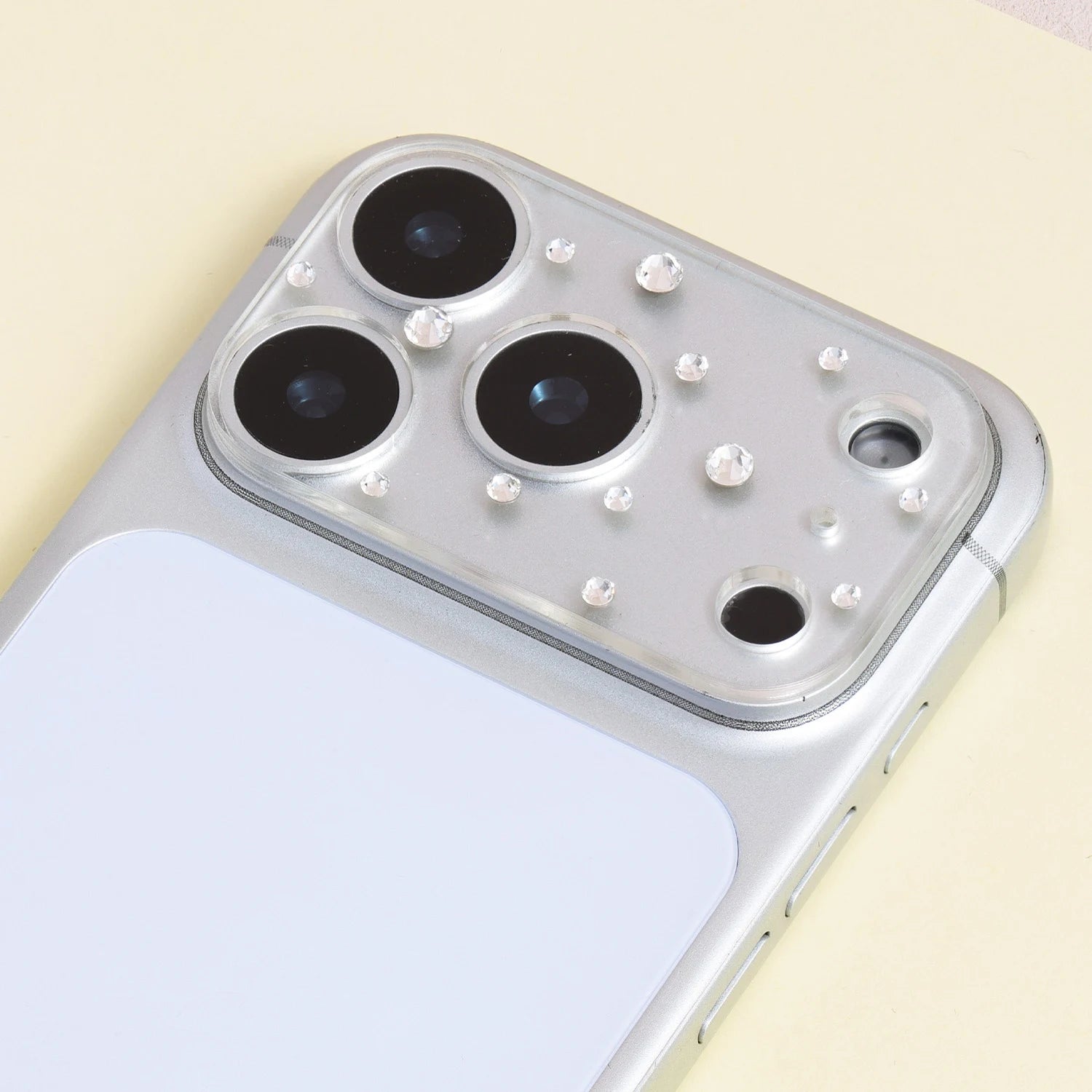 Silver crystal acrylic camera lens protector installed on a silver iPhone, showing delicate rhinestone accents around the camera area.