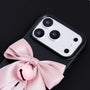 Close-up of a matte black phone case with an oversized blush pink satin bow and glossy pink bell accent beneath the camera frame.