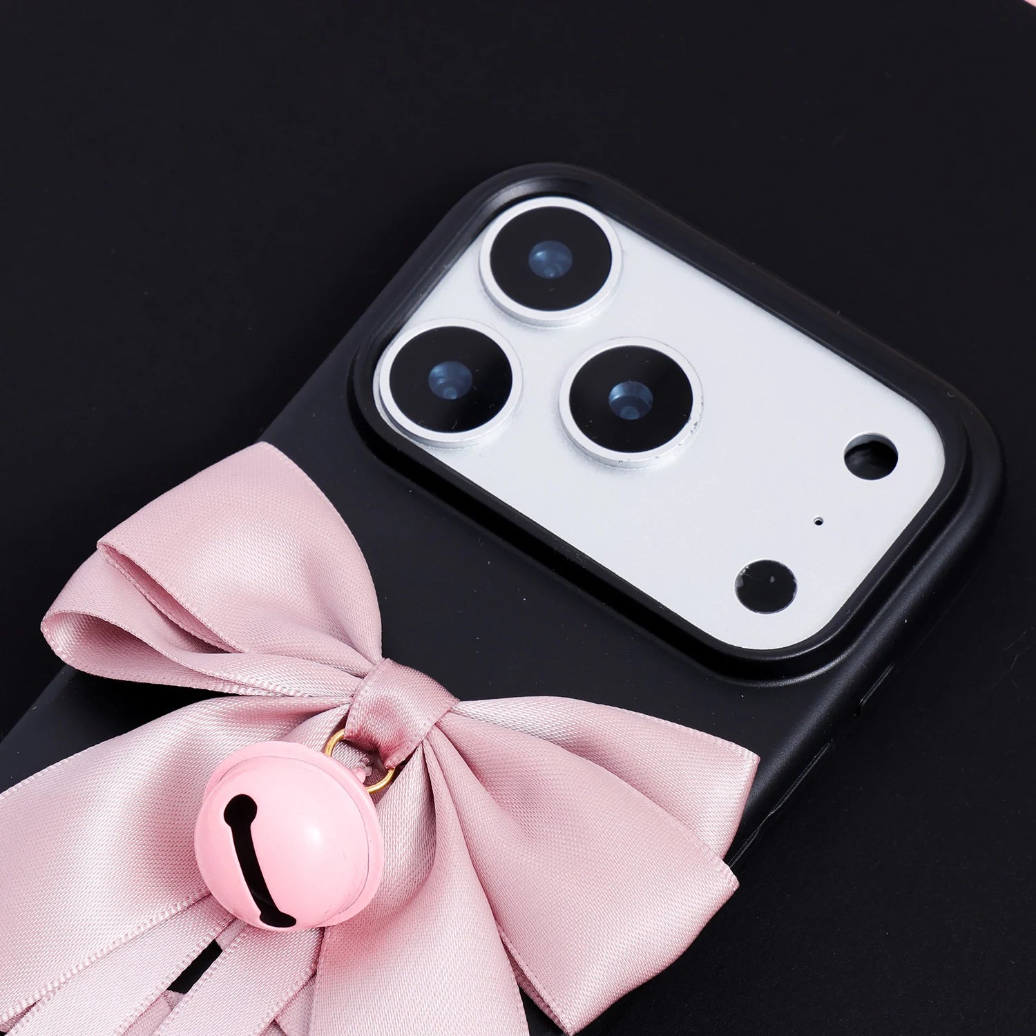 Close-up of a matte black phone case with an oversized blush pink satin bow and glossy pink bell accent beneath the camera frame.