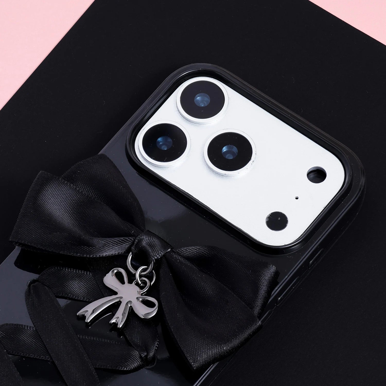 Close-up of a black handmade ribbon phone case showing the satin bow, silver pendant charm, glossy dark surface, and raised camera surround.