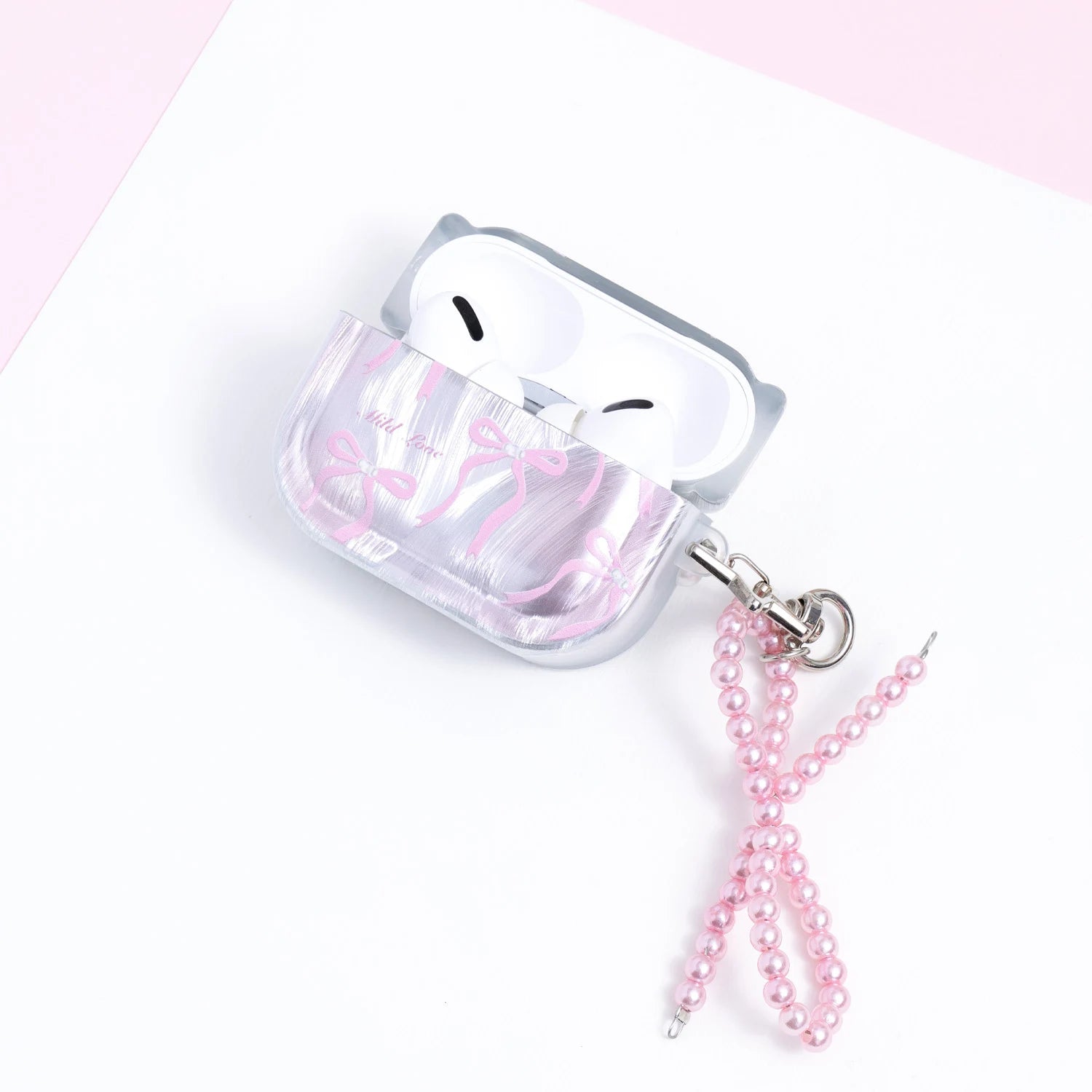 Open silver AirPods case with white AirPods inside, featuring pink bow motifs and a detachable pink pearl-bead charm at the side.
