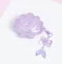 Closed lilac shell AirPods case with a glossy translucent finish, pearl and starfish detailing, and a dangling bow-and-mermaid-tail charm.