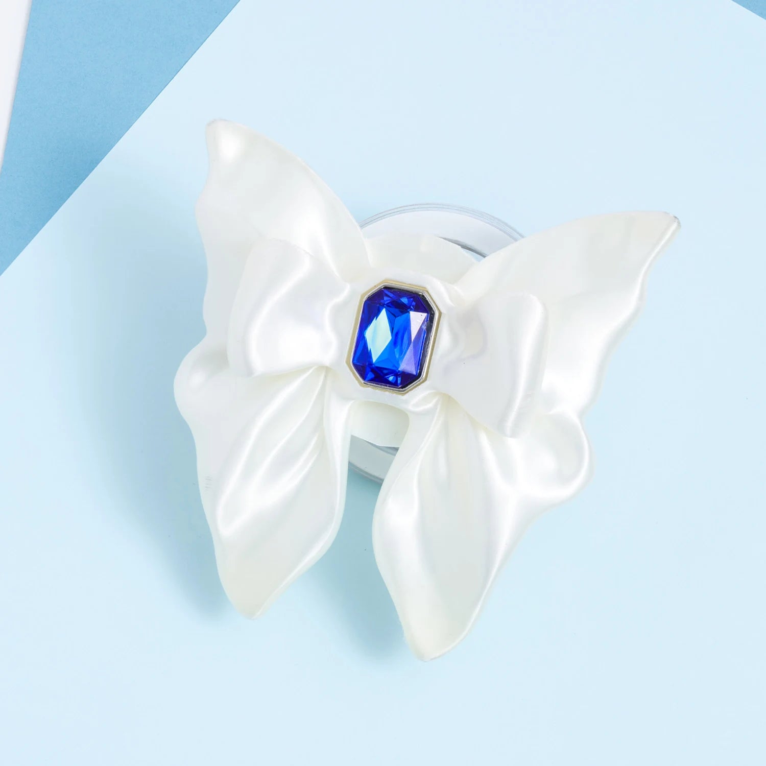 Front view of a white 3D-printed bow phone stand with smooth sculptural folds and a faceted blue jewel centerpiece on a pale blue background.
