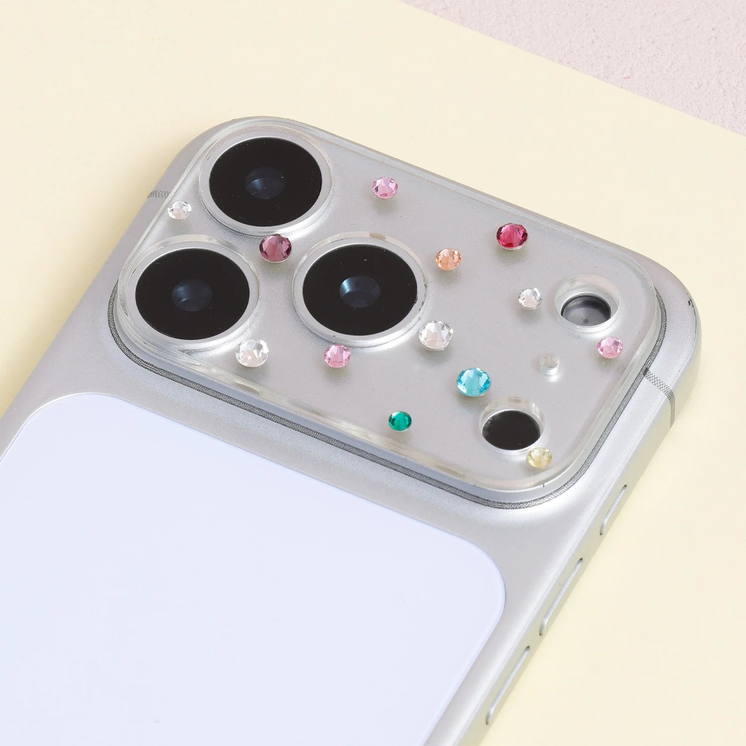 Multicolor crystal acrylic camera lens protector installed on a silver iPhone, showing jewel-like accents around the camera area.