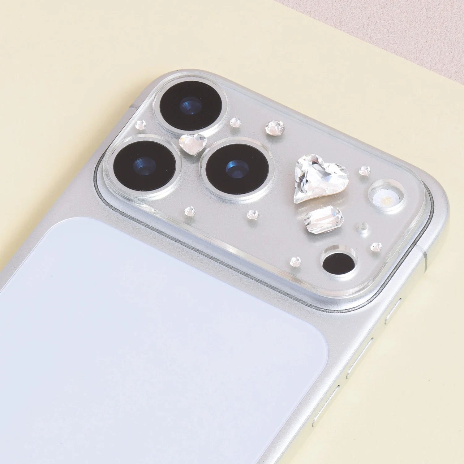 Heart crystal acrylic camera lens protector installed on a silver iPhone, showing a large clear heart gem and decorative accent stones around the camera area.
