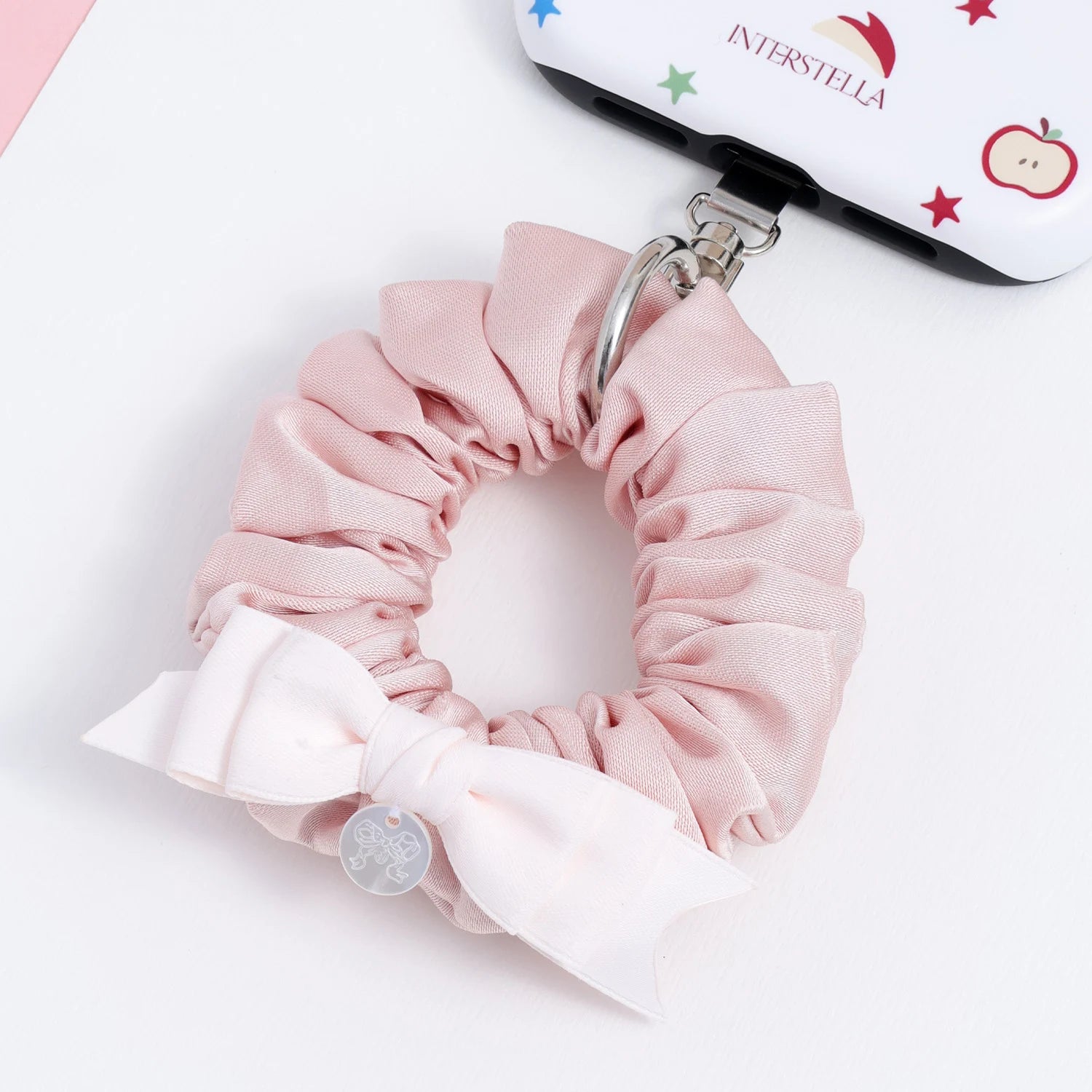 Blush pink phone hanging ring attached to a phone case, featuring a gathered fabric ring, ivory bow detail, and polished silver hardware.