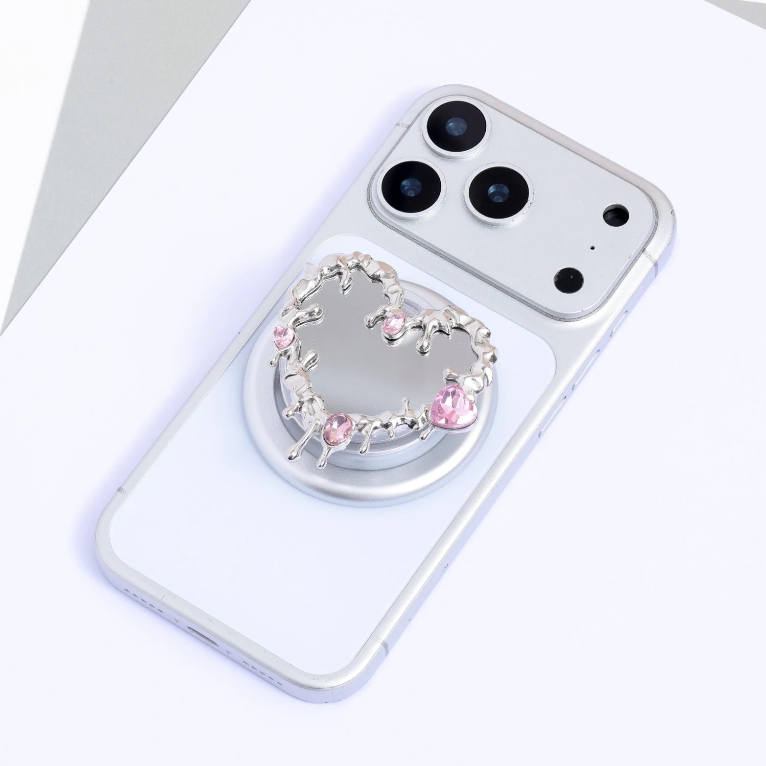 Heart-shaped silver-tone mirror phone stand with blush pink crystal accents attached to a silver phone on a circular stand base.