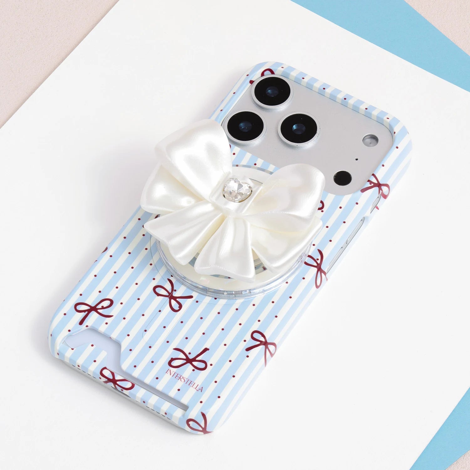 Ivory white jewel bow phone grip stand mounted on a blue-and-white striped phone case with red ribbon motifs.