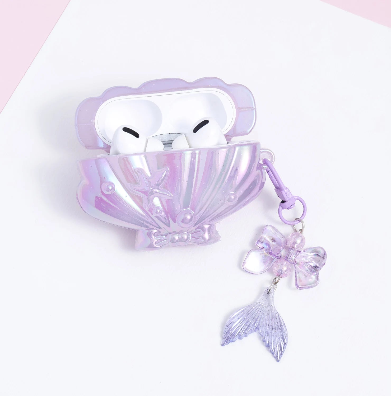Open lilac shell-shaped AirPods case with white AirPods inside, featuring an iridescent finish and a dangling translucent mermaid-tail charm.
