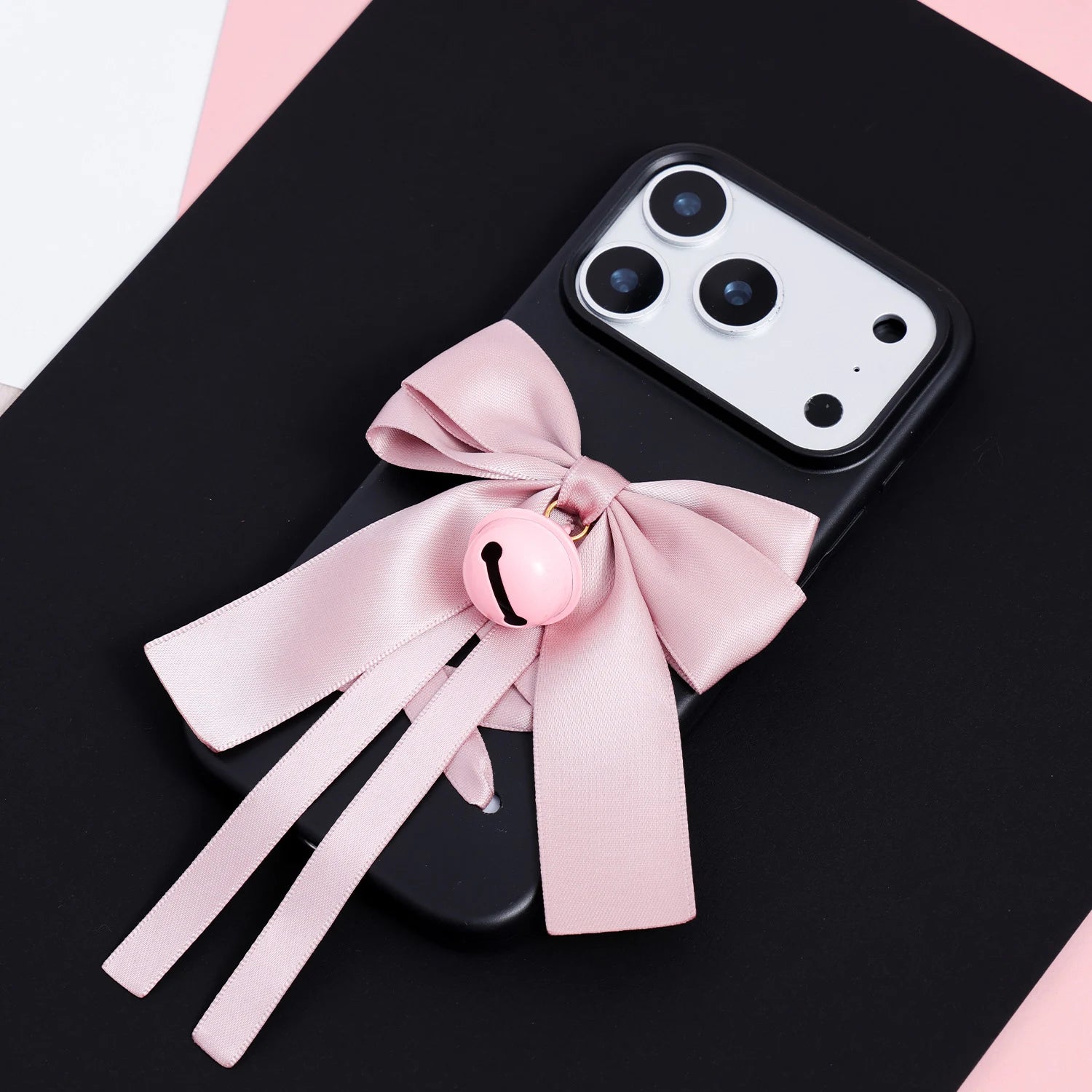 Full view of a matte black phone case with a large blush pink ribbon bow, long ribbon tails, and central bell charm on a dark background.