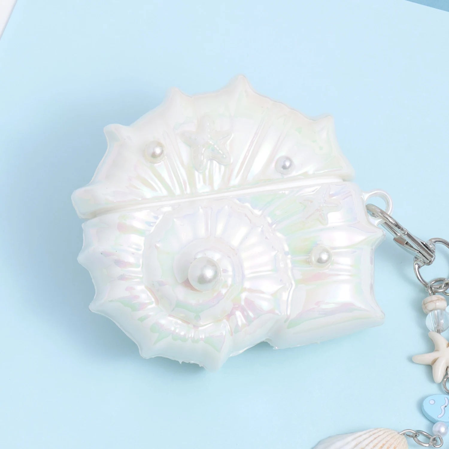 Close-up of an opaline conch-shaped AirPods case showing the spiral shell motif, pearl accents, glossy nacre-like finish, and starfish detailing.