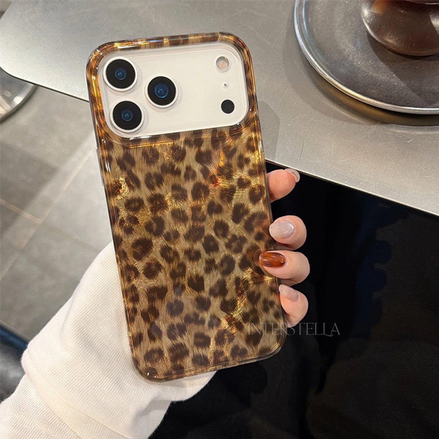 Close-up of a glossy petite leopard phone case laid on a metallic surface, highlighting the reflective finish, warm brown tones, and raised camera protection.