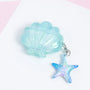 Closed aqua shell AirPods case with a glossy translucent surface and a dangling blue-iridescent starfish charm.
