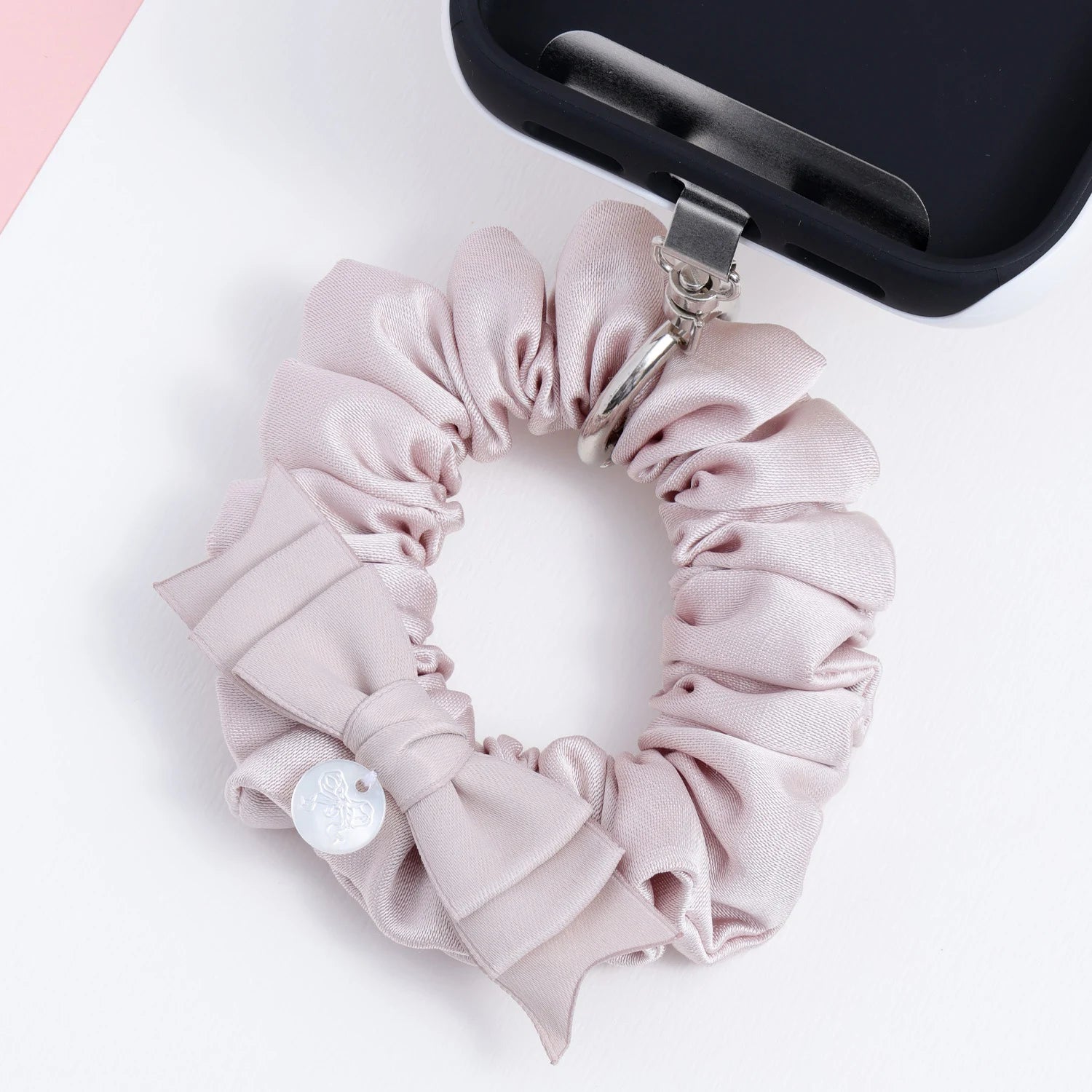 Dusty rose phone hanging ring attached to a black phone case, featuring a tonal bow, gathered satin-like fabric, and polished metal ring hardware.