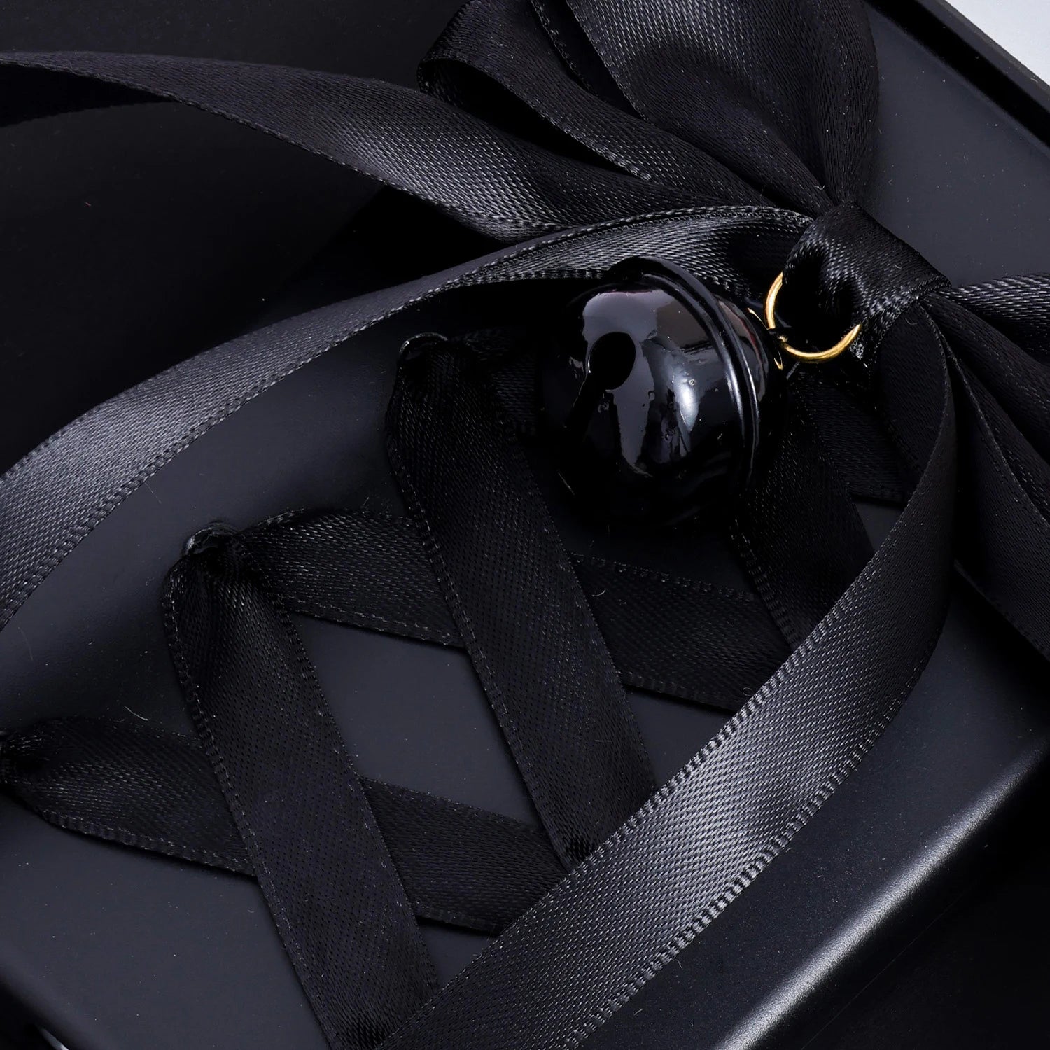 Detailed close-up of a black satin bow and glossy black bell charm on a matte black phone case with ribbon styling across the back.