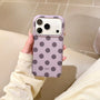 Hand holding a lilac phone case with charcoal grey polka dots and raised camera surround in a warm indoor setting.