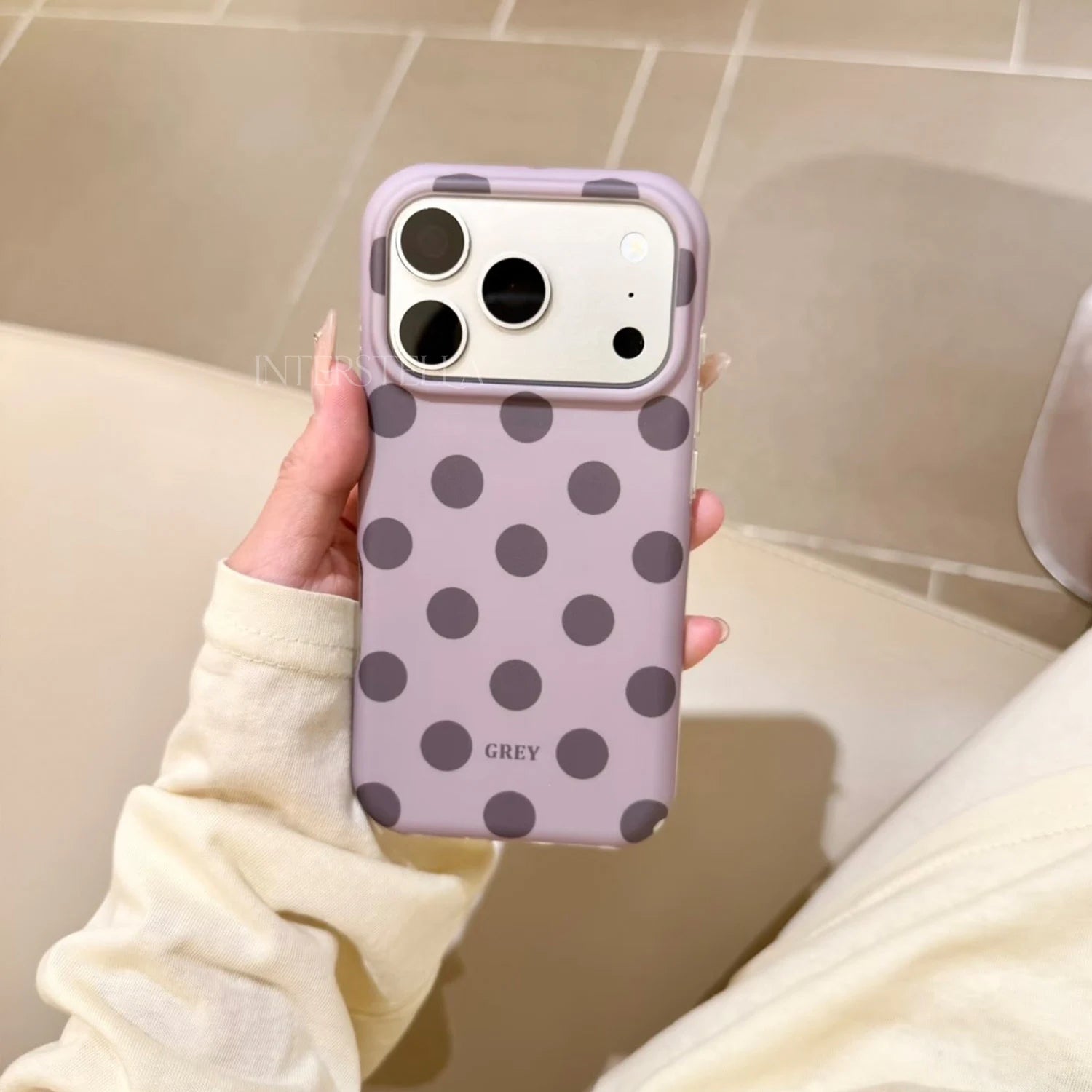 Hand holding a lilac phone case with charcoal grey polka dots and raised camera surround in a warm indoor setting.