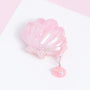 Closed pink shell AirPods case with a glossy blush iridescent finish and a dangling miniature shell-and-pearl charm.