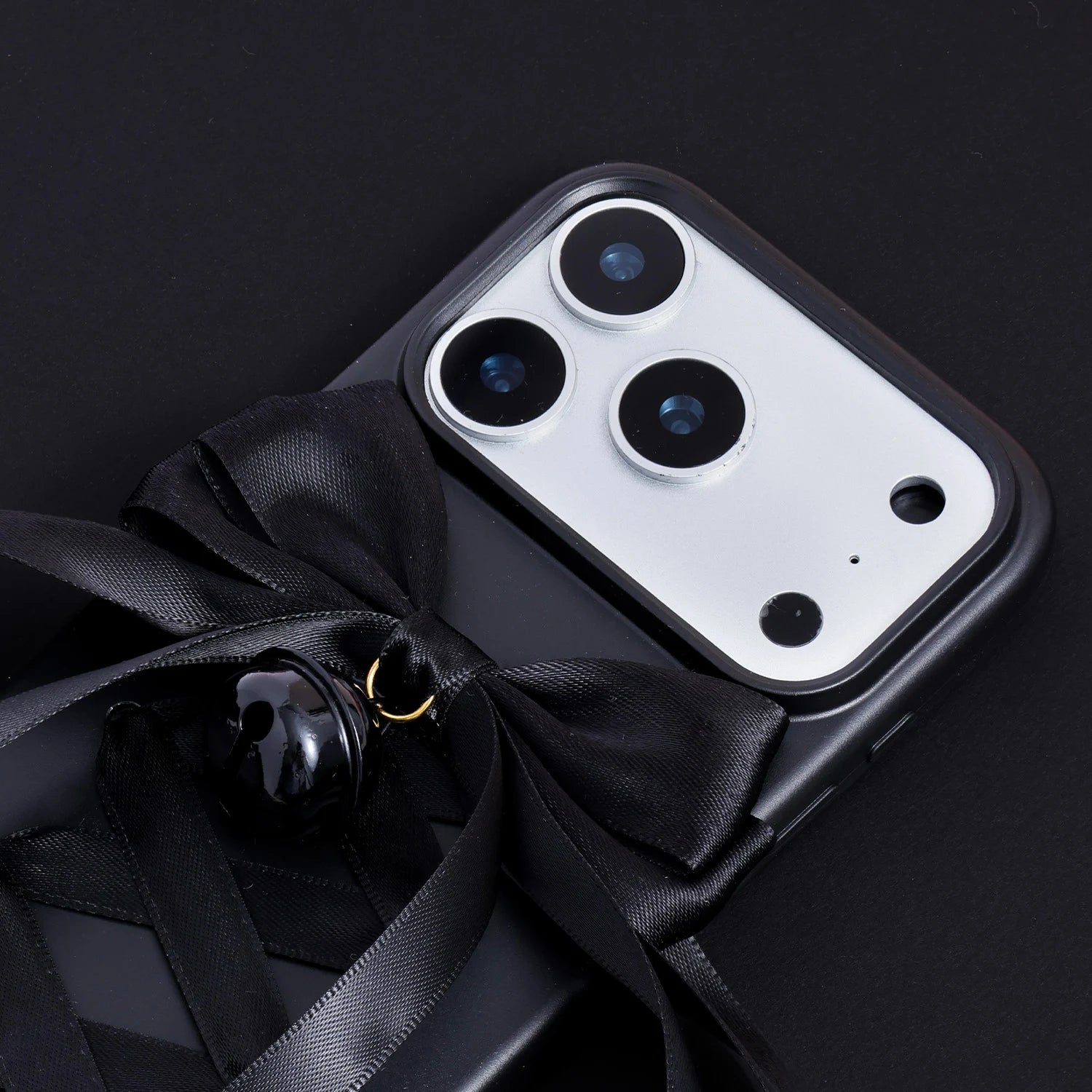 Close-up of a matte black phone case showing the black satin bow, glossy black bell accent, and raised camera surround.