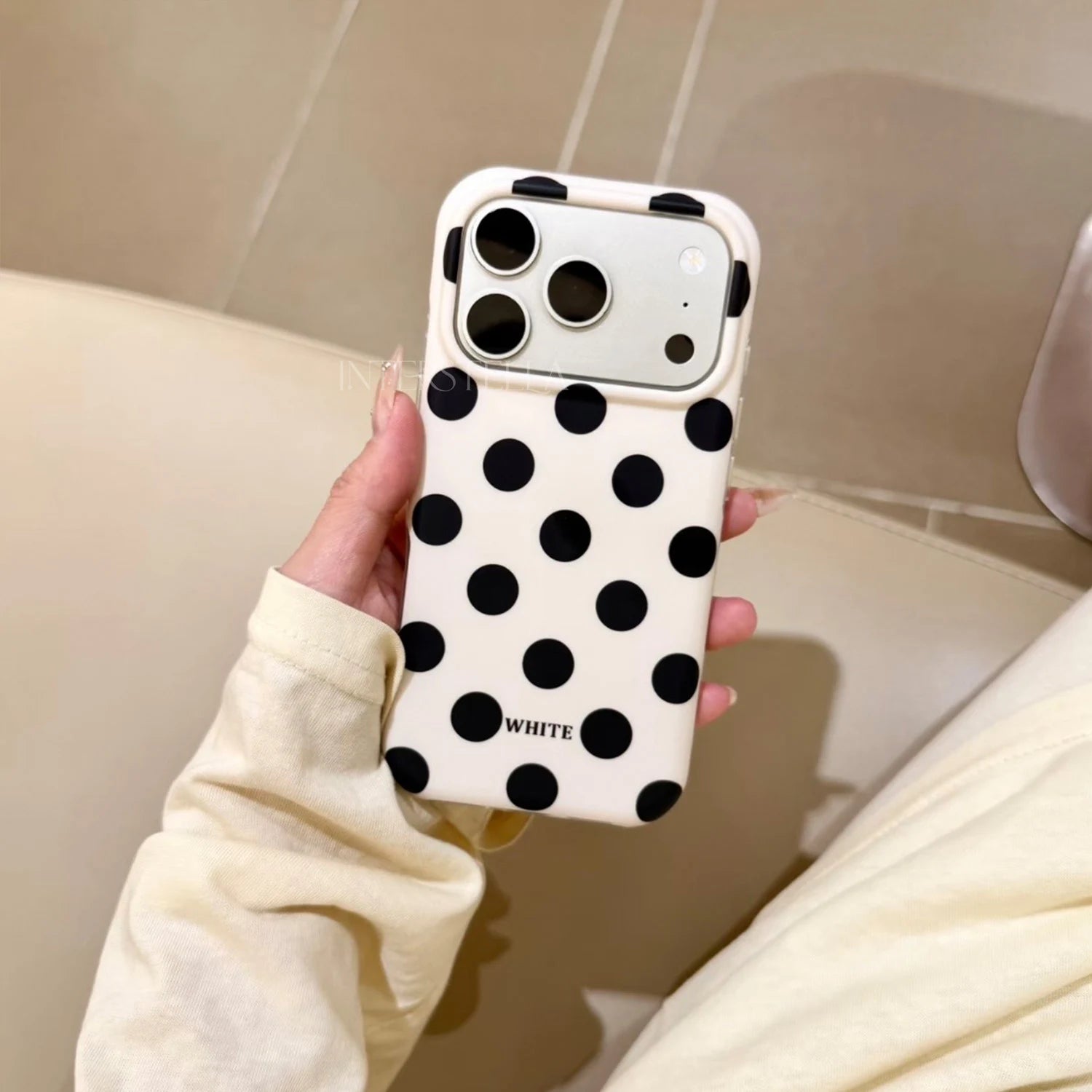 Hand holding an ivory phone case with bold black polka dots and raised camera surround in a warm indoor setting.
